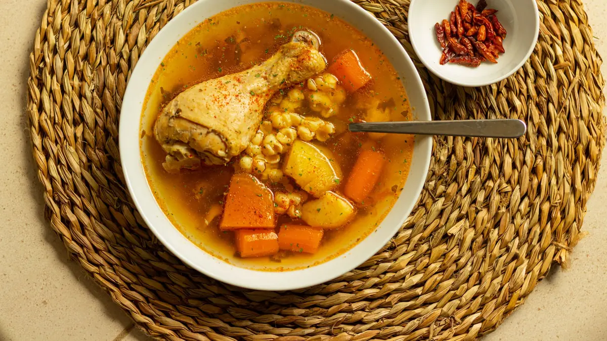Hvid dyb tallerken med klar hønsekødssuppe, en kyllingelår, gulerod, kartoffel og majskerner, set ovenfra på en flettet dækkeserviet
