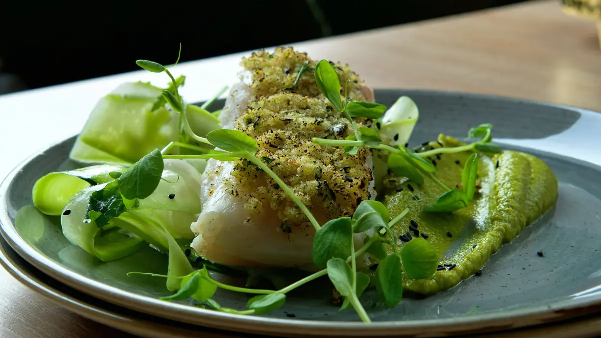 Torskefilet med sprød urteskorpe anrettet på grøn ærtepuré med tynde grøntsagsbånd og friske ærteskud