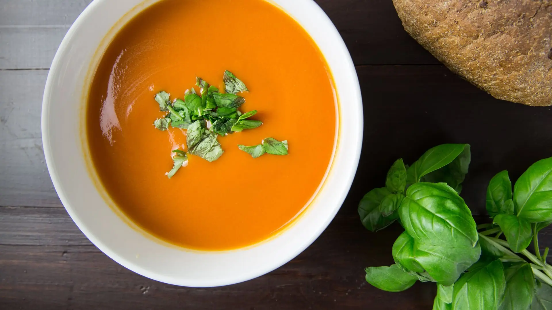 Hvid skål med rød tomatsuppe set oppefra, drysset med frisk basilikum, ved siden af et rustikt brød på mørkt træbord
