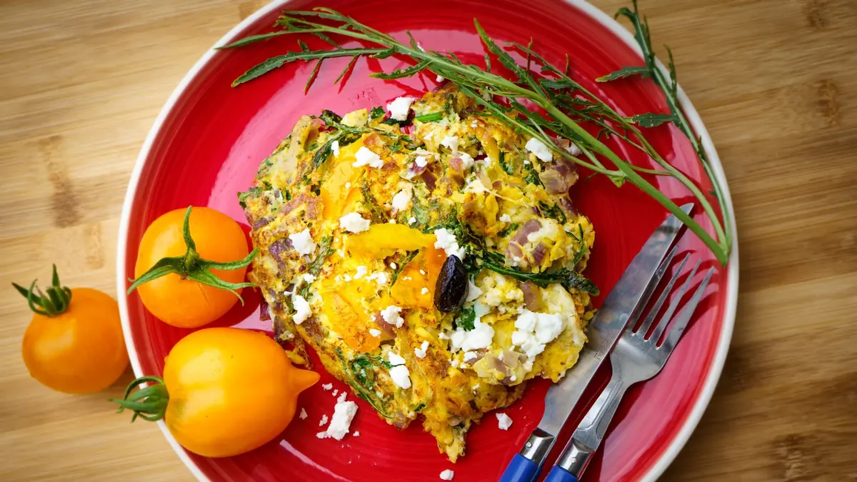 Frittata af æg med spinat, feta og rødløg på en rød tallerken med gule tomater ved siden af