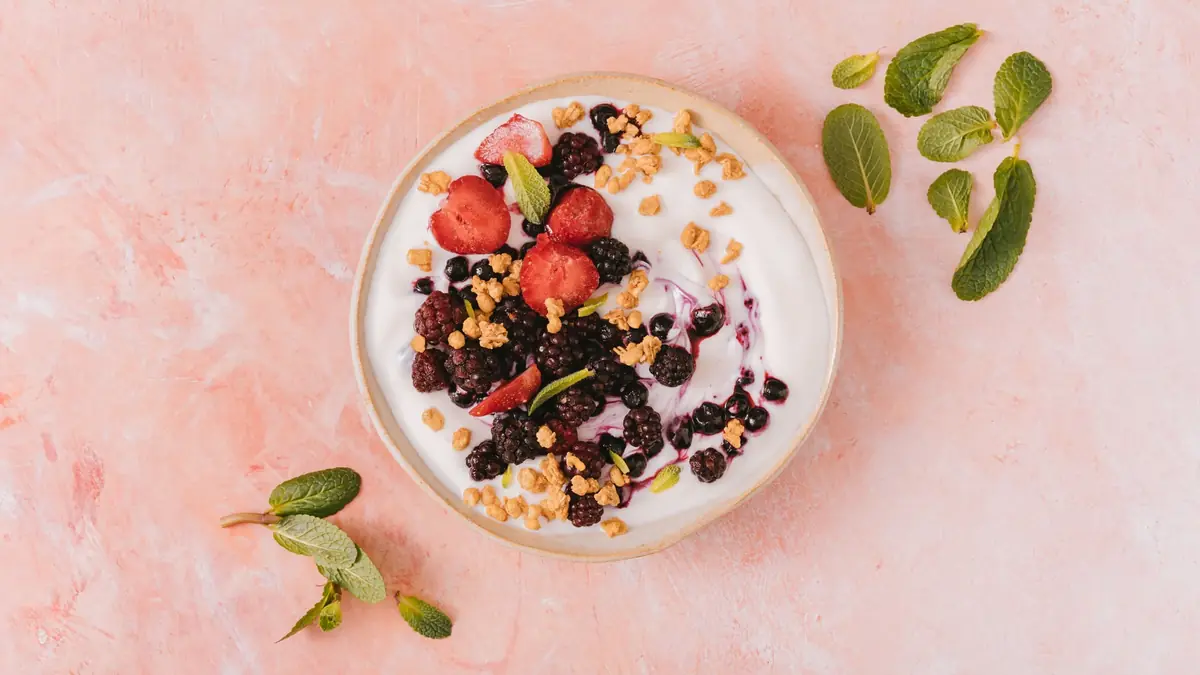 Skål med lys yoghurt toppet med brombær, blåbær og jordbær samt sprød mysli, set ovenfra på en lyserød flade med mynteblade ved siden af