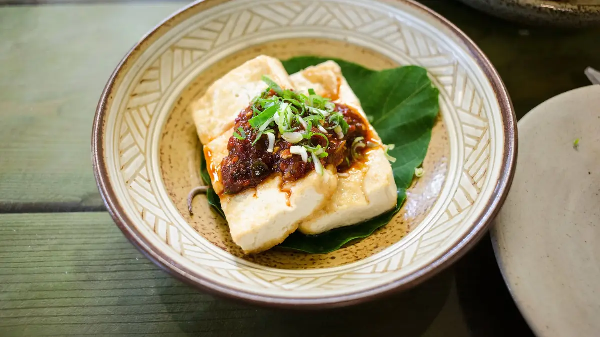 To skiver tofu med en klat mørkerød chilisauce og fintsnittede forårsløg, serveret på et grønt blad i en mønstret skål