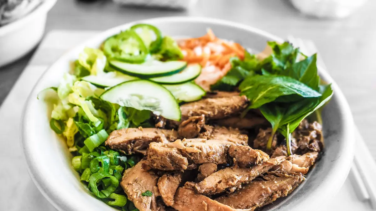 Skål med skiveskåret kylling, agurk, gulerod og salat med spisepinde ved siden af