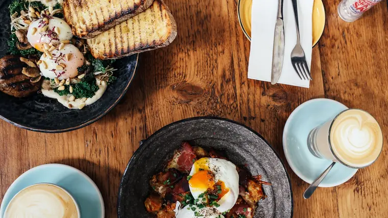 Brunchbord set ovenfra med pocherede æg på grønkål og svampe, grillet brød, en pande med bacon og æg samt to caffè latte på et træbord