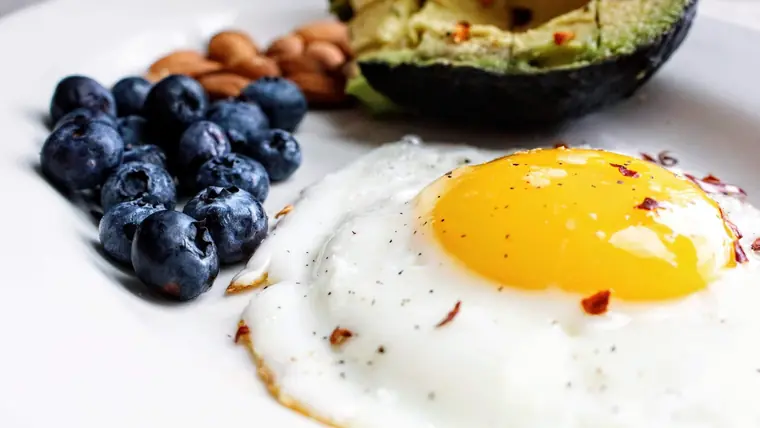 Spejlæg med løbende blomme, en dynge blåbær, mandler og en halv avocado på en hvid tallerken