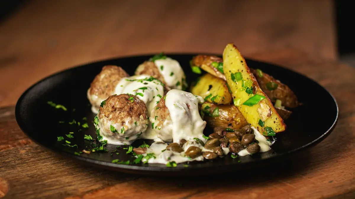 Frikadeller i cremet sauce anrettet på en sort tallerken sammen med ovnbagte kartoffelbåde