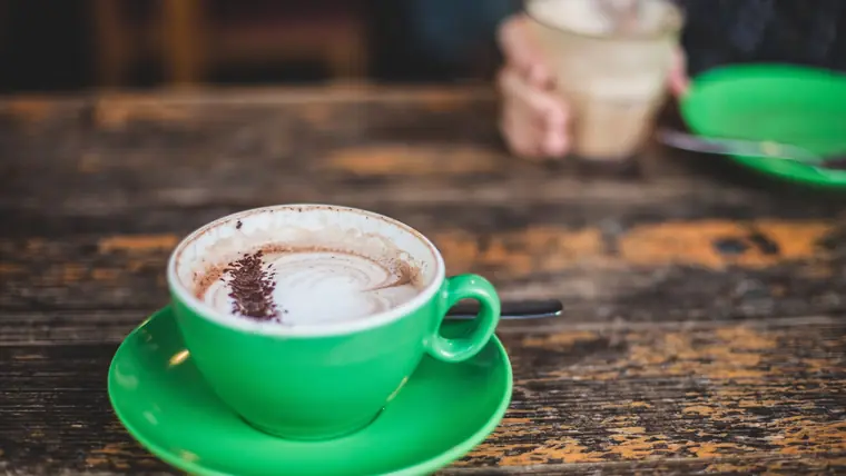 Grøn kaffekop med mælkeskum på et slidt træbord, med en hånd der holder endnu en kop i baggrunden