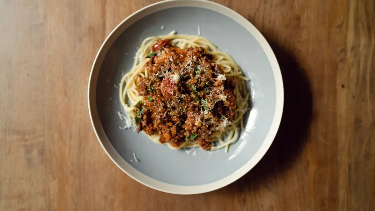 Spaghetti med kødsovs set oppefra, drysset med revet parmesan, på et mørkt træbord