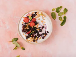 Skål med lys yoghurt toppet med brombær, blåbær og jordbær samt sprød mysli, set ovenfra på en lyserød flade med mynteblade ved siden af