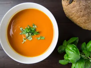 Hvid skål med rød tomatsuppe set oppefra, drysset med frisk basilikum, ved siden af et rustikt brød på mørkt træbord