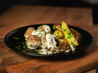 Frikadeller i cremet sauce anrettet på en sort tallerken sammen med ovnbagte kartoffelbåde