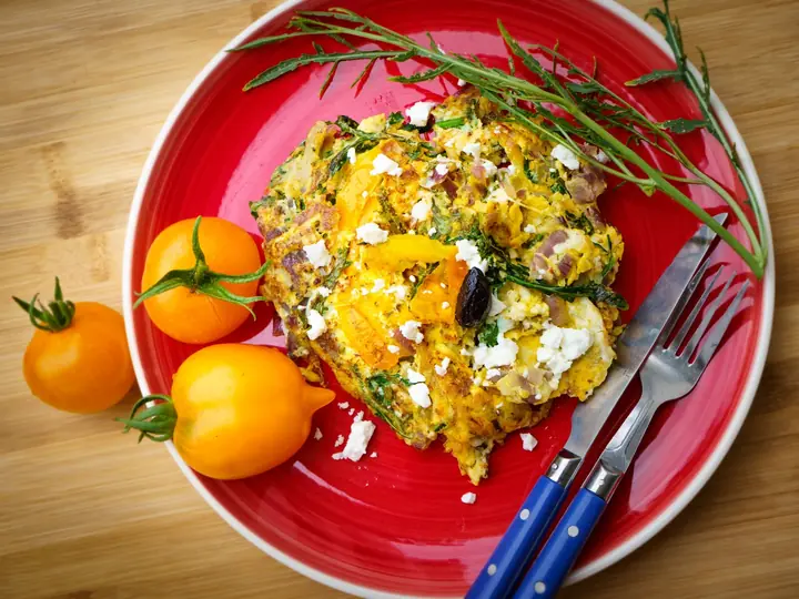 Frittata af æg med spinat, feta og rødløg på en rød tallerken med gule tomater ved siden af