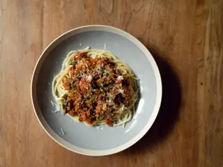 Spaghetti med kødsovs set oppefra, drysset med revet parmesan, på et mørkt træbord