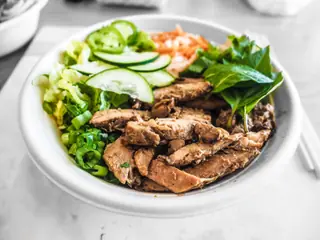 Skål med skiveskåret kylling, agurk, gulerod og salat med spisepinde ved siden af