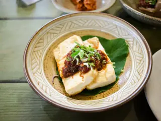 To skiver tofu med en klat mørkerød chilisauce og fintsnittede forårsløg, serveret på et grønt blad i en mønstret skål