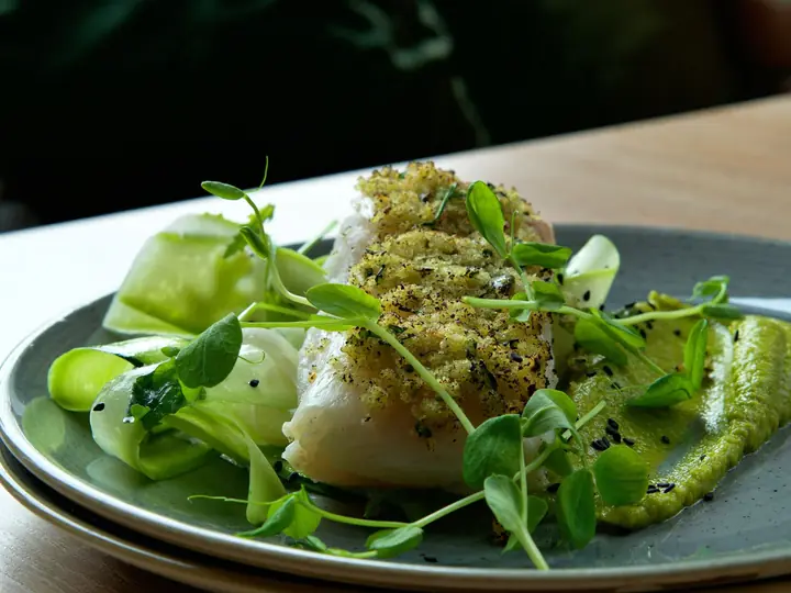 Torskefilet med sprød urteskorpe anrettet på grøn ærtepuré med tynde grøntsagsbånd og friske ærteskud
