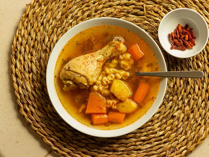Hvid dyb tallerken med klar hønsekødssuppe, en kyllingelår, gulerod, kartoffel og majskerner, set ovenfra på en flettet dækkeserviet