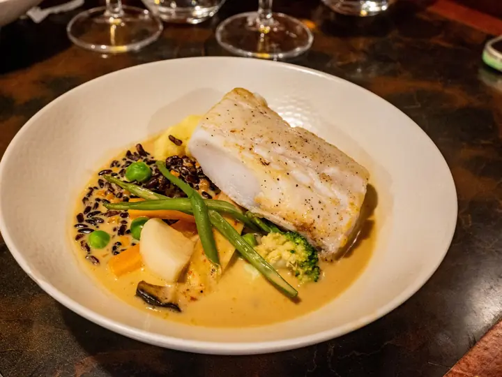 Hvid skål med et stykke torsk, gulerod, broccoli, ærter og bønner i en lys sauce